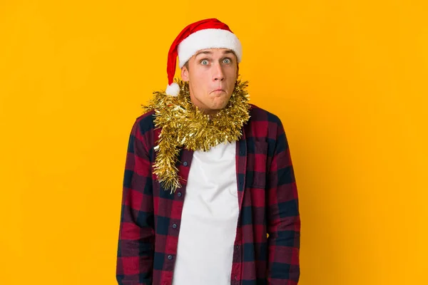 若いですCaucasian男とともにクリスマス帽子保持していますプレゼント絶縁上の黄色の背景Shrugs肩とオープン目混乱 — ストック写真
