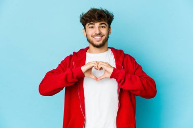 Mavi arka plandaki genç Arap adam gülümsüyor ve elleriyle kalp şeklini gösteriyor..