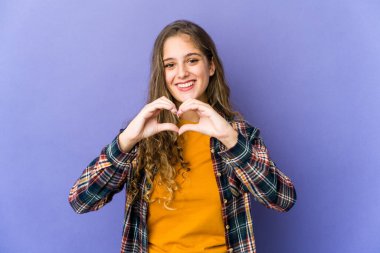 Beyaz tenli hoş bir kadın gülümsüyor ve elleriyle kalp şekli gösteriyor..