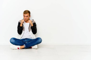 Yerde oturan genç beyaz bir adam kameraya yumruğunu gösteriyor, agresif bir yüz ifadesi..