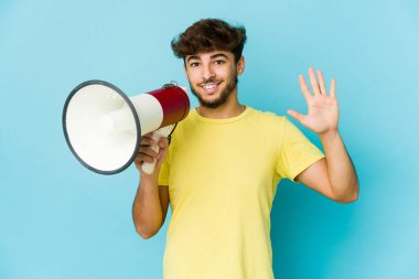 Genç Arap adam elinde megafonla gülümsüyor beş numarayı parmaklarıyla gösteriyor..