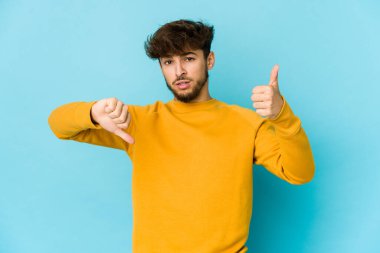 Mavi arka plandaki genç Arap adam baş parmağını kaldırıyor ve baş parmağıyla işaret ediyor. Zor seçim kavramı.