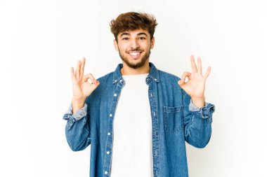 Beyaz arka plandaki genç Arap adam neşeli ve kendinden emin, iyi bir jest yapıyor..