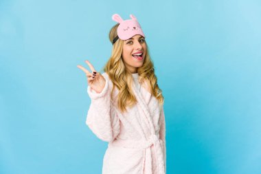 Genç sarışın beyaz kadın pijama giyiyor neşeli ve kaygısız parmaklarıyla barış sembolü gösteriyor..