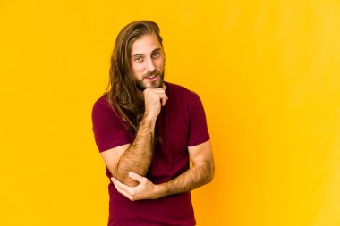 Uzun saçlı genç adam gülümsüyor mutlu ve kendinden emin, çenesine elle dokunuyor..