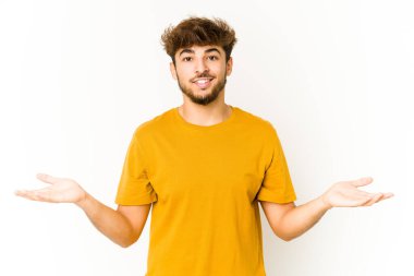 Beyaz arka plandaki genç Arap adam kollarıyla ölçeği yapıyor, mutlu ve kendinden emin hissediyor..