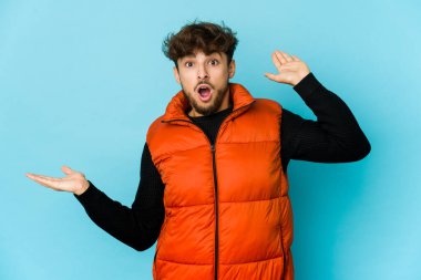 Mavi arka plandaki genç Arap 'ın elinde avuç içi fotokopi alanı var. Şaşırmış ve mutlu..