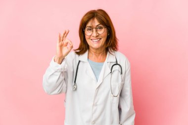 Orta yaşlı beyaz doktor kadın neşeli ve kendinden emin bir şekilde iyi bir jest yapıyor..