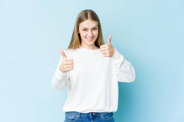 Genç sarışın kadın mavi arka planda izole edilmiş. Baş parmağını kaldırıyor, gülümsüyor ve kendinden emin..