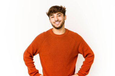 Beyaz arka plandaki genç Arap adam kendine güveniyor. Ellerini kalçasından ayırmıyor..