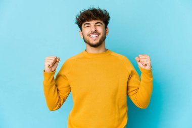 Mavi arka plandaki genç Arap adam zaferi, tutkuyu ve coşkuyu, mutlu ifadeyi kutluyor..