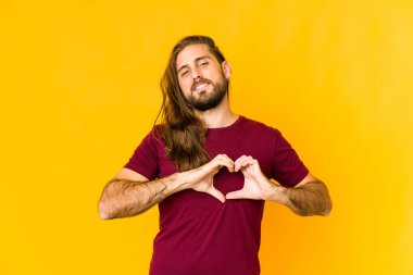 Uzun saçlı genç adam gülümsüyor ve elleriyle kalp şeklini gösteriyor..