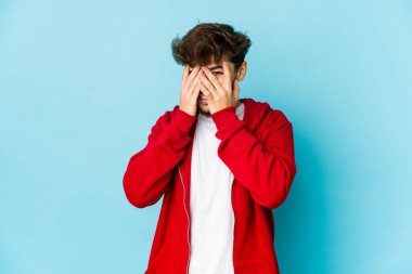 Mavi arka plandaki genç Arap adam kameraya parmaklarıyla göz kırpıyor, yüzünü kapatmaktan utanıyor..