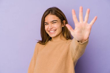 Mor arka planda genç, sıska beyaz kız gülümseyerek beş numarayı parmaklarıyla gösteriyor..