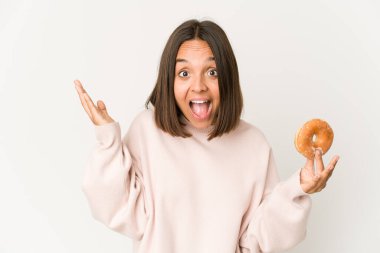 Young hispanic woman eating a doughnut receiving a pleasant surprise, excited and raising hands.