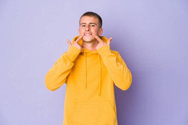 Beyaz tenli yakışıklı adam gülümsüyor, parmaklarıyla ağzını işaret ediyor..