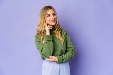 Genç beyaz sarışın kadın gülümsüyor mutlu ve kendinden emin, elleriyle çenesine dokunuyor..