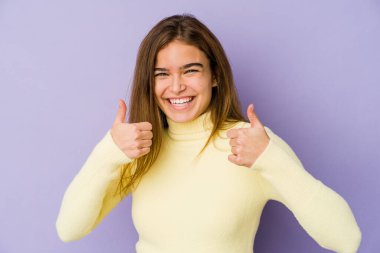Mor arka planda genç, sıska, beyaz bir kız gülümsüyor ve başparmağını kaldırıyor.