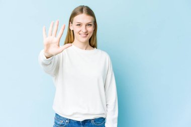 Mavi arka planda izole edilmiş genç sarışın kadın gülümseyerek beş numarayı parmaklarıyla gösteriyor..