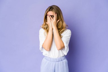 Genç, beyaz, sarışın kadın parmaklarının arasından korkmuş ve gergin bir şekilde göz kırpıyor..