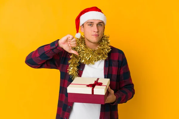 クリスマスの帽子をかぶった若い白人男性が黄色の背景に孤立したプレゼントを持っており 嫌なジェスチャー 親指を下に示しています 意見の相違 — ストック写真