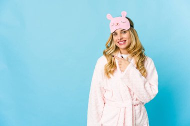 Pijama giyen, gülümseyen ve boş alanda bir şeyler gösteren sarışın beyaz bir kadın..