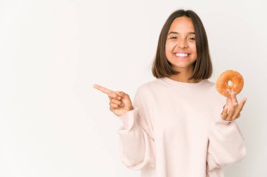 Young hispanic woman eating a doughnut smiling and pointing aside, showing something at blank space.