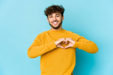 Mavi arka plandaki genç Arap adam gülümsüyor ve elleriyle kalp şeklini gösteriyor..