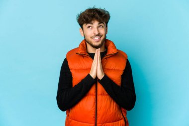 Mavi arka planda el ele tutuşan genç bir Arap. Ağız kenarında dua ediyor. Kendine güveniyor..