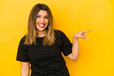 Sarı arka planda izole edilmiş genç Hintli kadın işaret parmağıyla işaret ederek gülümsüyor..