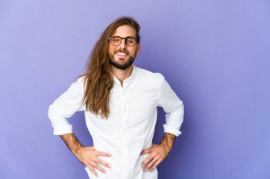 Uzun saçlı genç adam kendine güveniyor gibi görünüyor. Ellerini kalçasında tutuyor..