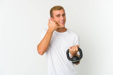 Beyaz arka planda izole edilmiş kettlebell tutan beyaz tenli genç bir adam parmaklarıyla bir cep telefonu hareketi gösteriyor..