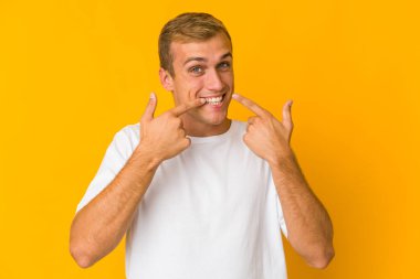 Beyaz tenli yakışıklı adam gülümsüyor, parmaklarıyla ağzını işaret ediyor..