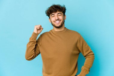 Mavi arka plandaki genç Arap adam zaferi, tutkuyu ve coşkuyu, mutlu ifadeyi kutluyor..