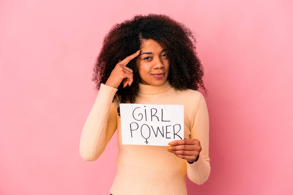 Young african american curly woman holding a girl power message on a placard pointing temple with finger, thinking, focused on a task.