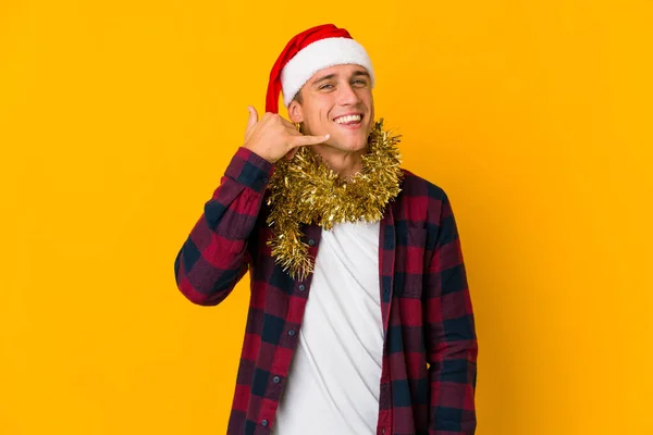 头戴圣诞礼帽的年轻高加索人拿着一个黄色背景的礼物 手握着一个手指头打手机的手势 — 图库照片