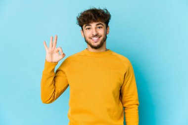 Mavi arka plandaki genç Arap adam neşeli ve kendinden emin, iyi bir jest yapıyor..