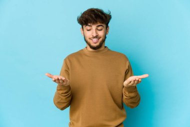 Mavi arka planda genç bir Arap elinde avuç içi bir şeyle kameraya sunuyor..