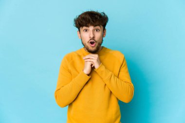 Mavi arka plandaki genç Arap adam şans için dua ediyor, şaşırmış ve ağzını açıyor..