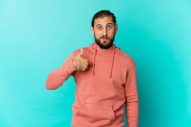 Uzun saçlı genç adam parmaklarıyla işaret ederek şaşırmış görünüyor, geniş bir gülümsemeyle..