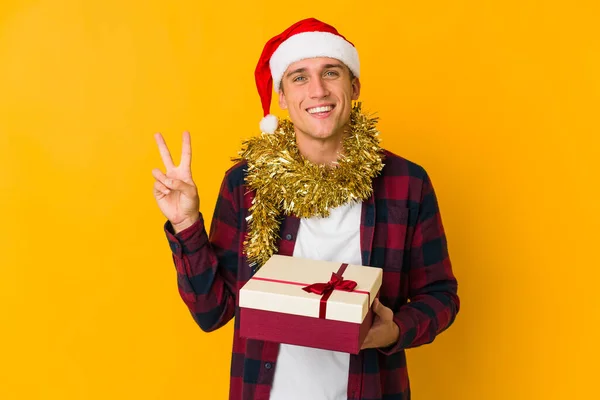 クリスマスの帽子をかぶった若い白人男性が黄色の背景に孤立したプレゼントを持っています喜びと屈託のない指で平和のシンボルを示す — ストック写真