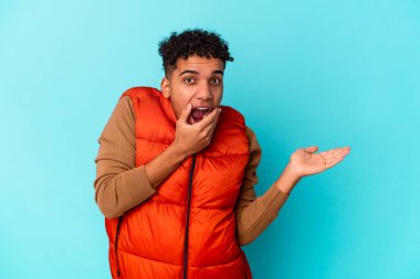 Mavi üzerine izole edilmiş genç Afro-Amerikalı kıvırcık adam avuç içinde fotokopi alanı tutuyor, el üstünde tutuyor. Şaşırmış ve mutlu..