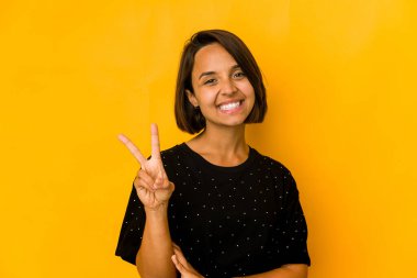 Sarı üzerine izole edilmiş genç İspanyol kadın zafer işareti gösteriyor ve gülümsüyor..