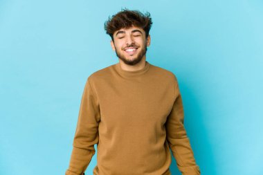 Mavi arka plandaki genç Arap adam gülüyor ve gözlerini kapatıyor, rahatlamış ve mutlu hissediyor..