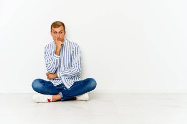 Genç, beyaz bir adam yerde oturuyor. Şüpheli ve şüpheci bir ifadeyle yan bakıyor..