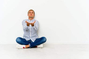 Genç, beyaz bir adam yerde oturuyor. Dudaklarını kıvırıyor ve avuç içlerine hava öpücüğü yolluyor..