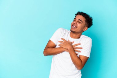 Genç Afro-Amerikan kıvırcık adamı mavi üzerine izole edilmiş gülüyor, ellerini kalbe koyuyor, mutluluk kavramını koruyor..