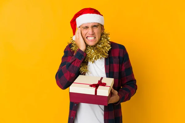 若いですCaucasian男とともにクリスマス帽子保持していますプレゼント隔離された上に黄色の背景カバー耳で手 — ストック写真