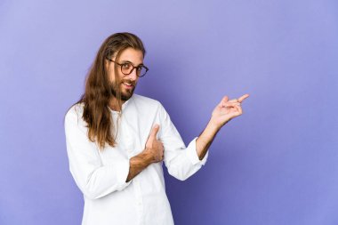 Uzun saçlı genç adam işaret parmağıyla işaret ederek gülümsüyor..