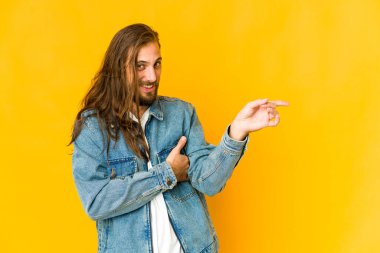 Uzun saçlı genç adam işaret parmağıyla işaret ederek gülümsüyor..